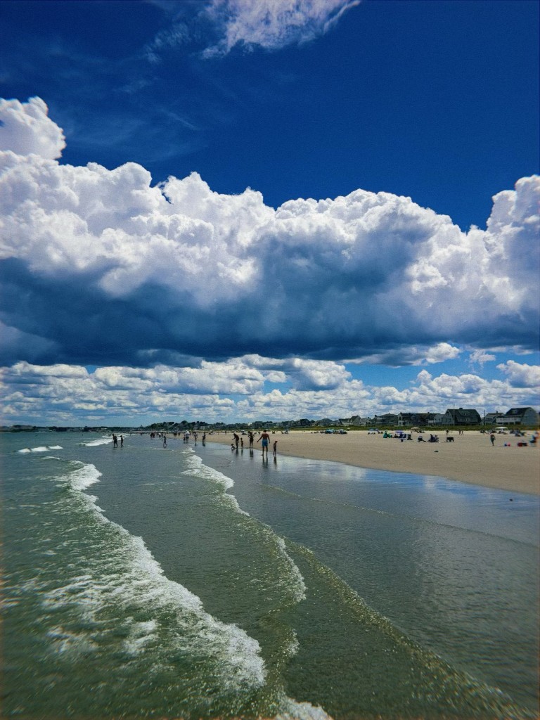 Wide beach under towering summer clouds, waves rolling into shore with people wading in the shallows
